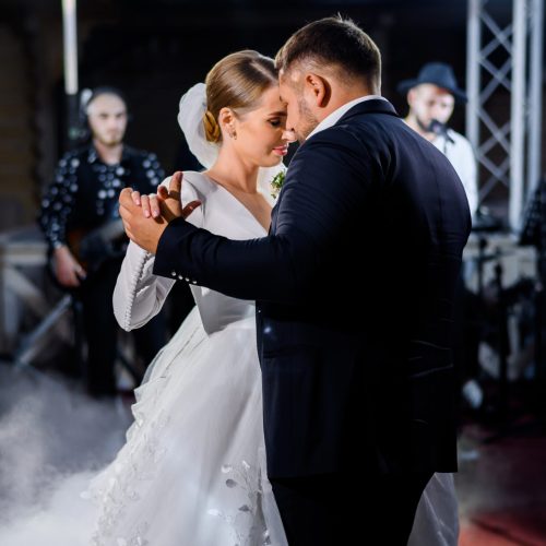 Side view of loving bride in wedding puffy dress and veil with groom in suit, dancing first dance in decorated hall with smoke while band playing on scene on background